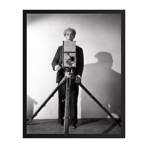 Puede incluir: Una fotografía en blanco y negro que muestra a una persona con gorra plana y traje oscuro, de pie detrás de una cámara de cine vintage tipo caja montada sobre un trípode de madera alto de tres patas, situada a la altura de los ojos.