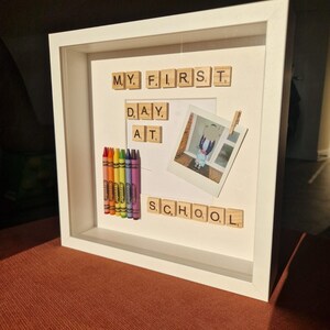 Könnte beinhalten: Weißer Schattenrahmen mit dem Text "MY FIRST DAY AT SCHOOL", der mit Holzbuchstaben geschrieben wurde. Enthält ein Foto, eine Wäscheklammer und eine Reihe bunter Buntstifte.
