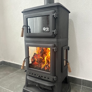 Può includere: Stufa a legna in ghisa nera con una porta in vetro e un fuoco che brucia all'interno. La stufa ha un design decorativo e una maniglia in legno. La stufa è etichettata "Alp Soba".