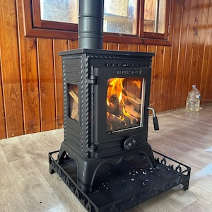 May include: A black cast iron wood-burning stove with a glass door and a black metal base. The stove is lit and has flames visible inside. The stove is in front of a wooden wall.