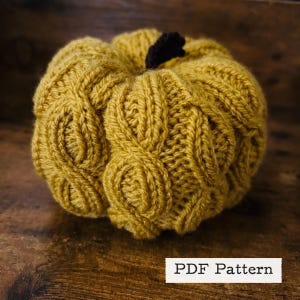 Könnte beinhalten: Ein gestrickter Kürbis in senfgelber Farbe mit einem Zopfmuster. Der Kürbis liegt auf einer Holzoberfläche. PDF-Muster.