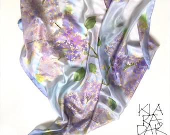 Könnte beinhalten: Ein hellblauer Seidenschal mit einem Aquarell-Design aus lila und goldenen Fliederblüten. Der Schal ist gefaltet und auf einem weißen Hintergrund drapiert.