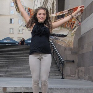May include: A woman wearing a black tank top and light gray jeans jumps in the air while holding a colorful scarf. She is wearing black flats and has curly brown hair.