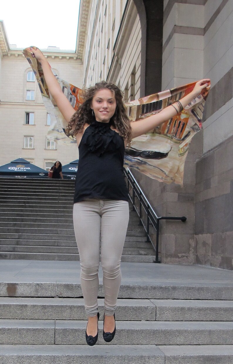 May include: A woman wearing a black tank top and light gray jeans jumps in the air while holding a colorful scarf. She is wearing black flats and has curly brown hair.