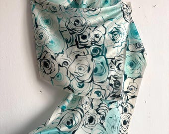 Puede incluir: Bufanda de seda con un estampado de rosas en tonos turquesa, negro y blanco. La bufanda está colgada de una percha, mostrando la textura fluida y el diseño. Las rosas son detalladas y artísticas.