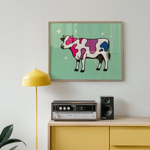 Pode incluir: Uma impressão emoldurada com uma vaca de desenho animado com manchas rosa, roxas e azuis em um fundo verde claro. A vaca está situada em um fundo de estrelas brancas brilhantes. A impressão é exibida acima de um sistema de som e um alto-falante preto.