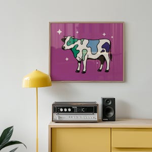 May include: A framed art print featuring a cartoon cow with colorful patches of green, blue, and purple on a white body. The cow is set against a purple background with white star accents. The print is in a wooden frame.