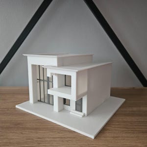 Peut inclure: Maquette architecturale blanche d'une maison moderne avec un toit plat, de grandes fenêtres et un balcon. Le modèle repose sur une base blanche et est exposé sur une étagère en bois. La maquette est une représentation miniature du design contemporain.
