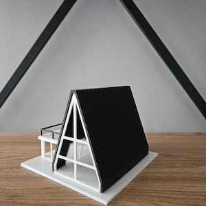 May include: A miniature A-frame house model with a black roof and white frame. The model sits on a white base and features a small balcony. The house is a modern architectural design.