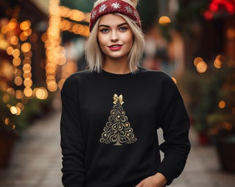May include: Black sweatshirt featuring a gold and brown Christmas tree design with a bow at the top. The model is wearing a red and white headband with a snowflake pattern. The background is blurred with warm, golden lights.