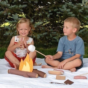 May include: Two children are sitting by a pretend campfire, toasting marshmallows and sausages. The campfire is made of felt and has a yellow flame. There are also felt sausages and chocolate bars on the white blanket.