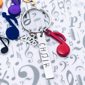 May include: A silver keychain with a rectangular charm that reads "CHOIR" and the year "2026". The keychain also features a red musical note charm and other colorful musical note charms.