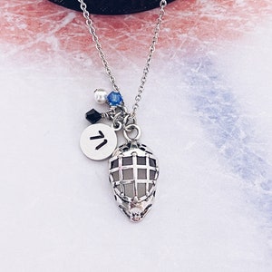 May include: A silver necklace with a hockey mask charm, a small round charm with the letter "V" on it, and three small beads. The beads are white, blue, and black.