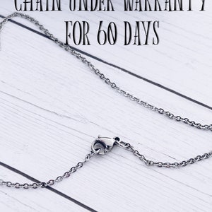 May include: A silver chain necklace with a lobster clasp. The chain is on a white wooden surface. The text "CHAIN UNDER WARRANTY FOR 60 DAYS" is visible above the chain.