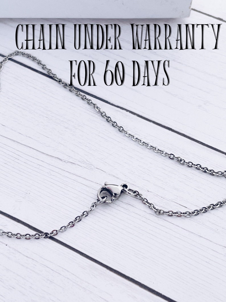May include: A silver chain necklace with a lobster clasp. The chain is on a white wooden surface. The text "CHAIN UNDER WARRANTY FOR 60 DAYS" is visible above the chain.