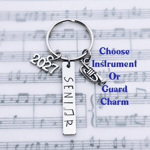 May include: A silver-toned metal keychain featuring a circular ring with three attached charms: a rectangular tag stamped with the word SENIOR, a charm displaying the number 2027, and a small trumpet-shaped charm. The text Choose Instrument Or Guard Charm is visible.