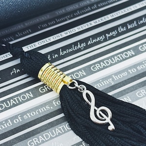May include: Black graduation tassel with a gold band and a silver treble clef charm. The background features text from a graduation card with quotes and the word "GRADUATION" repeated.