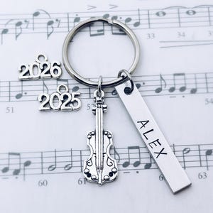 May include: A silver keychain with a violin charm, a rectangular tag with "ALEX" engraved, and charms with the numbers "2025" and "2026". The keychain is on a background of musical notes.
