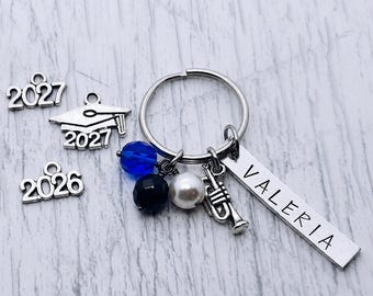 May include: A silver-toned metal keychain featuring a rectangular bar charm engraved with the name VALERIA, a trumpet charm, a white pearl bead, a black bead, and a blue bead. Additional loose charms include a graduation cap and the years 2026 and 2027.