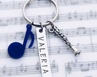 May include: A silver-toned metal keychain featuring three charms: a bright blue acrylic musical note, a rectangular silver metal tag engraved with the name VALERIA in capital letters, and a small silver-toned metal clarinet instrument charm.