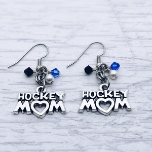 May include: Silver-toned hockey mom earrings. Each earring features the words "HOCKEY MOM" with a heart-shaped cutout. The earrings have a dangle design with black and blue beads and silver-toned hooks.