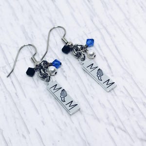 May include: Silver dangle earrings with the letters "MOM" and a winged design. The earrings have silver hooks, black and blue beads, and small white pearls. The earrings are displayed on a white and gray marble surface.