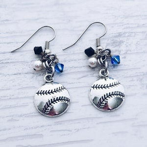 May include: A pair of baseball-themed earrings. Each earring features a silver-toned baseball charm with black stitching. The charms are accented with black and blue beads, and a white pearl. The earrings have silver-colored fish hook ear wires.