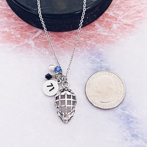 May include: A silver necklace with a hockey mask charm, a silver charm with the letter "T" and blue and white beads. The necklace is laying on a white surface with a silver US quarter for scale.
