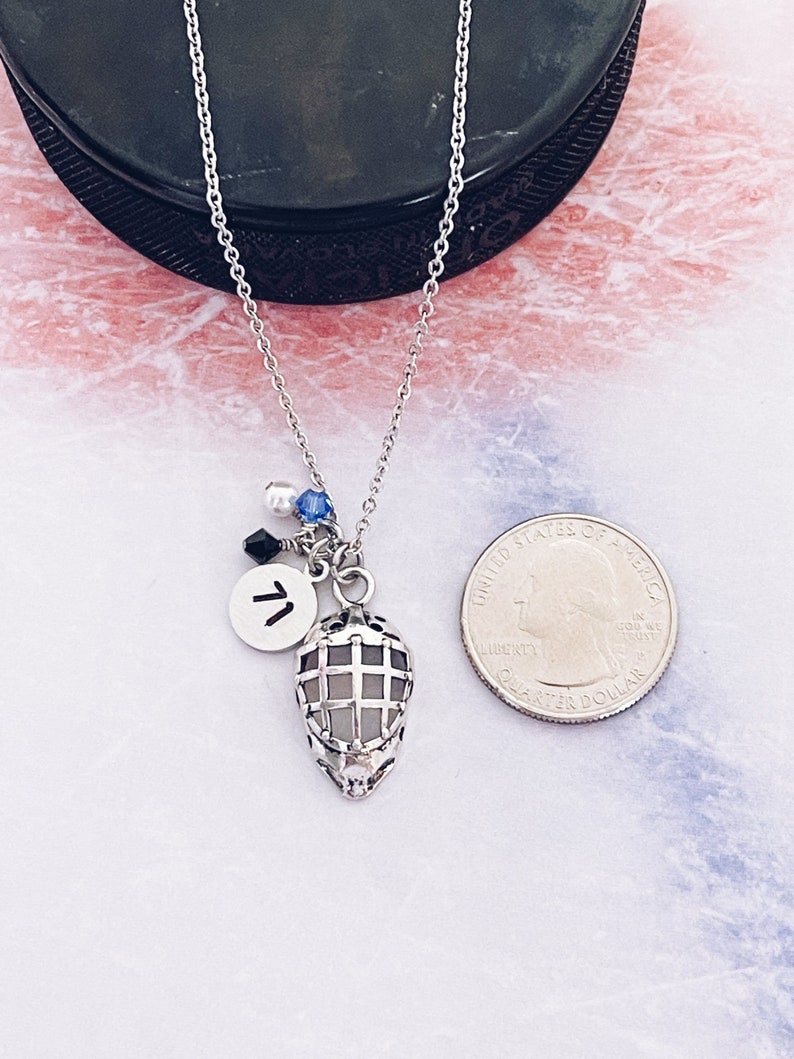 May include: A silver necklace with a hockey mask charm, a silver charm with the letter "T" and blue and white beads. The necklace is laying on a white surface with a silver US quarter for scale.