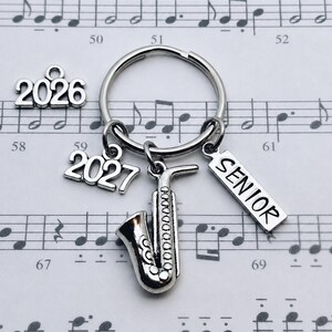 May include: A silver-toned metal keychain featuring a saxophone charm, a rectangular charm engraved with the word SENIOR, and a charm displaying the year 2027. An additional loose charm with the year 2026 is positioned to the left of the ring.