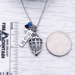 Puede incluir: Un collar de plata con un dije de m&aacute;scara de hockey, un dije de plata con la palabra "MOM" en &eacute;l y algunas peque&ntilde;as cuentas azules y negras. El collar est&aacute; al lado de una moneda de 25 centavos de d&oacute;lar estadounidense para referencia de tama&ntilde;o.