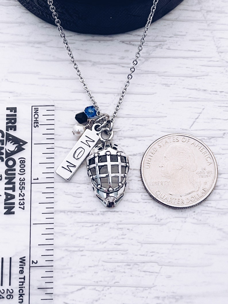 Puede incluir: Un collar de plata con un dije de m&aacute;scara de hockey, un dije de plata con la palabra "MOM" en &eacute;l y algunas peque&ntilde;as cuentas azules y negras. El collar est&aacute; al lado de una moneda de 25 centavos de d&oacute;lar estadounidense para referencia de tama&ntilde;o.