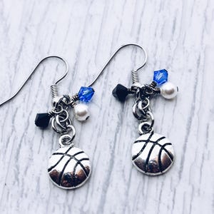 May include: Silver basketball earrings with black and blue beads and white pearls. The earrings have a hook closure and are designed with a basketball charm. The earrings are on a white and gray background.