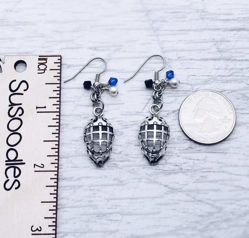 May include: Silver hockey mask earrings with blue and white beads. The earrings are about 1 inch long and are shown next to a ruler and a US quarter.