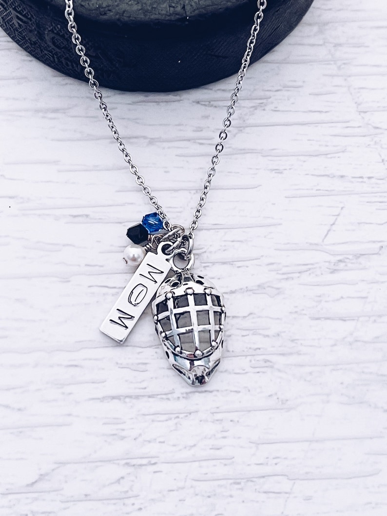 Puede incluir: Un collar de plata con un dije de m&aacute;scara de hockey, una etiqueta de plata que dice "MOM" y cuentas azules, negras y blancas.