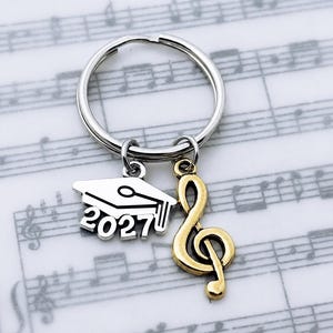 May include: A silver-toned metal keyring featuring two charms: a silver-colored graduation cap charm with the year 2027 embossed on the front and a gold-toned metal treble clef music note charm hanging from a circular split ring.