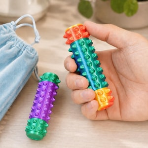 May include: A colorful sensory toy with a cylindrical shape and textured spikes. The toy is held in a hand, showcasing its rainbow gradient design. A smaller, similar toy in purple and green is also visible.