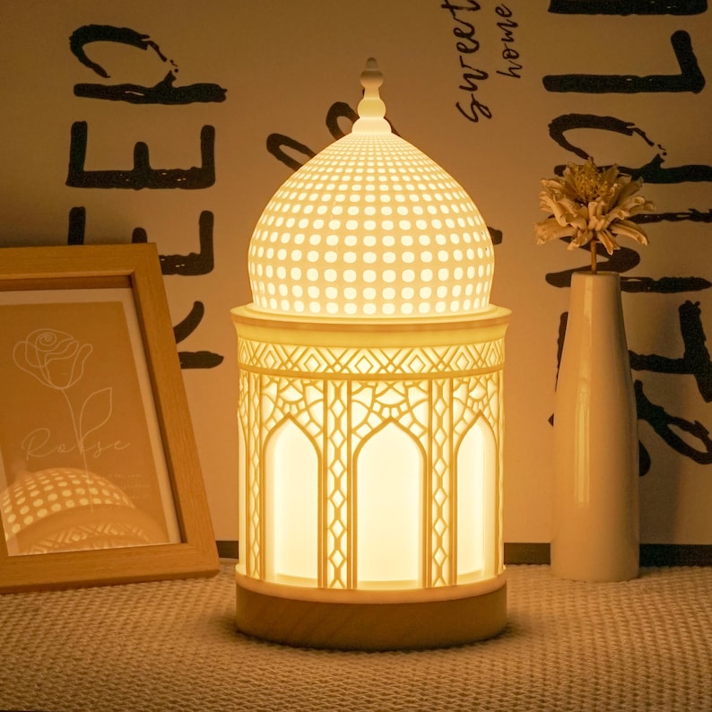 May include: White ceramic table lamp with a dome-shaped shade featuring a geometric pattern. The lamp base has a decorative, intricate design with arches and a warm white glow.