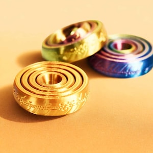 Op de afbeelding: Drie kleurrijke, metalen fidget spinners in goud, groen en blauw. De gouden spinner staat op de voorgrond, met gegraveerde tekst rond de rand. De andere twee spinners staan op de achtergrond, met een cirkelvormig ontwerp.