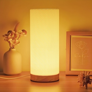 Puede incluir: Una lámpara de mesa cilíndrica con base de madera y pantalla texturizada de color blanquecino. La lámpara está encendida, emitiendo un brillo cálido. Un jarrón con flores y un cuadro enmarcado también están sobre la mesa. La lámpara mide unos 30 cm de alto.