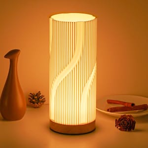 Puede incluir: Una lámpara de mesa cilíndrica que emite una luz cálida. La lámpara presenta un diseño acanalado vertical con un patrón fluido. La base es de madera. Otros elementos decorativos incluyen un jarrón, una piña, palitos de canela y una rosa.