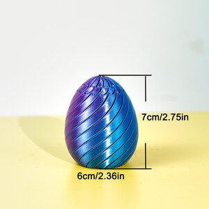 Op de afbeelding: Een decoratief eivormig object met een verloop van blauwe en paarse tinten. Het ei is 7 cm hoog en 6 cm breed, met een spiraalvormig ontwerp. Het object staat tegen een lichte achtergrond.