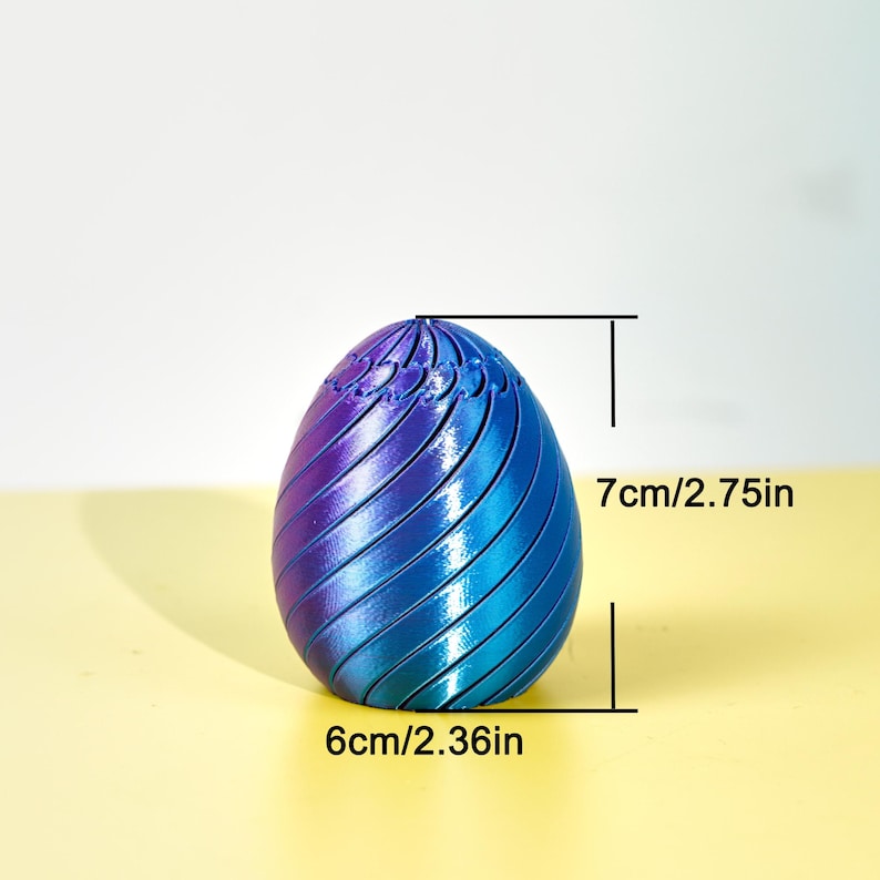 Op de afbeelding: Een decoratief eivormig object met een verloop van blauwe en paarse tinten. Het ei is 7 cm hoog en 6 cm breed, met een spiraalvormig ontwerp. Het object staat tegen een lichte achtergrond.