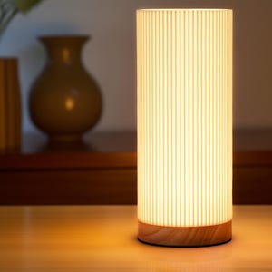May include: A white cylindrical lamp with a wood base. The lamp has vertical grooves and emits a warm, white light.