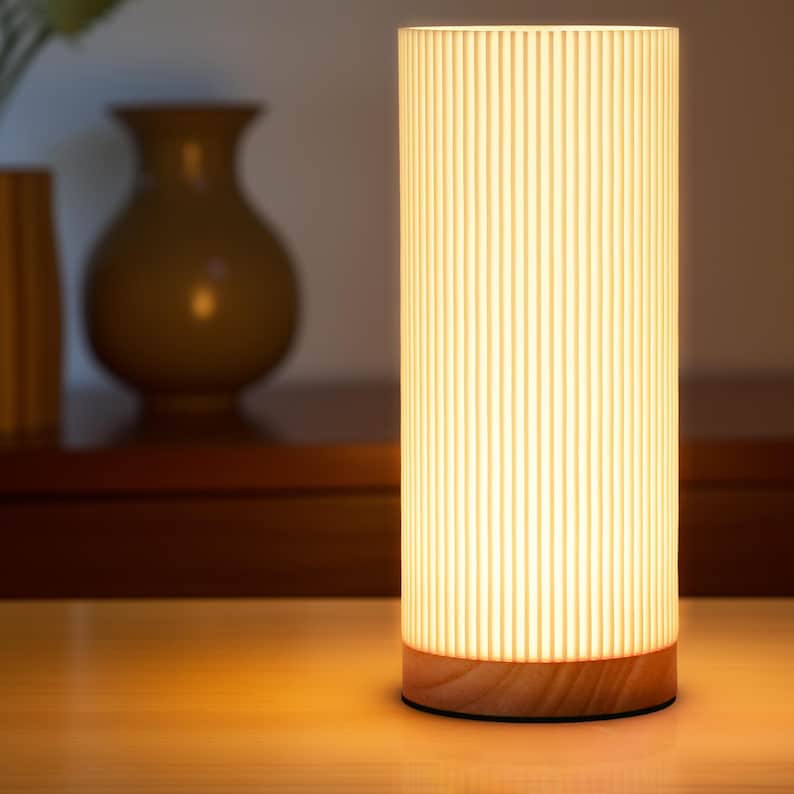 May include: A white cylindrical lamp with a wood base. The lamp has vertical grooves and emits a warm, white light.