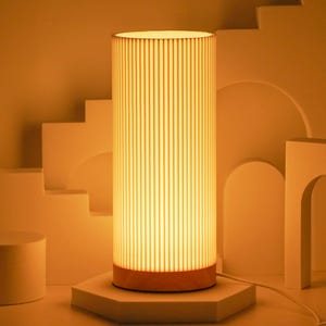 May include: A tall, cylindrical lamp with a white, ribbed shade and a wooden base. The lamp is lit and casting a warm glow.