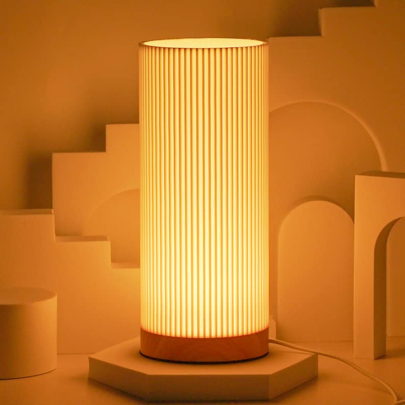 May include: A tall, cylindrical lamp with a white, ribbed shade and a wooden base. The lamp is lit and casting a warm glow.