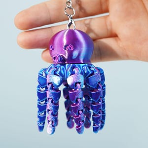 Puede incluir: Un llavero de pulpo impreso en 3D en tonos morados y azules. El pulpo tiene una cabeza redondeada con detalles de ojos y tentáculos articulados. El llavero está sujeto a un anillo de metal plateado.