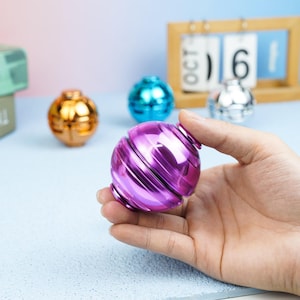 May include: A metallic purple spherical fidget toy with a smooth, reflective surface. The object features a segmented design with horizontal grooves. In the background, additional spherical fidget toys in gold, blue, and silver are visible.