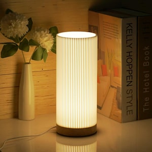 May include: A white cylindrical table lamp with a wood base and a white linen shade with vertical lines. The lamp is turned on and illuminating the room. The lamp is sitting on a white surface next to a stack of books with the text "Kelly Hoppen Style" and "The Hotel Book" visible.
